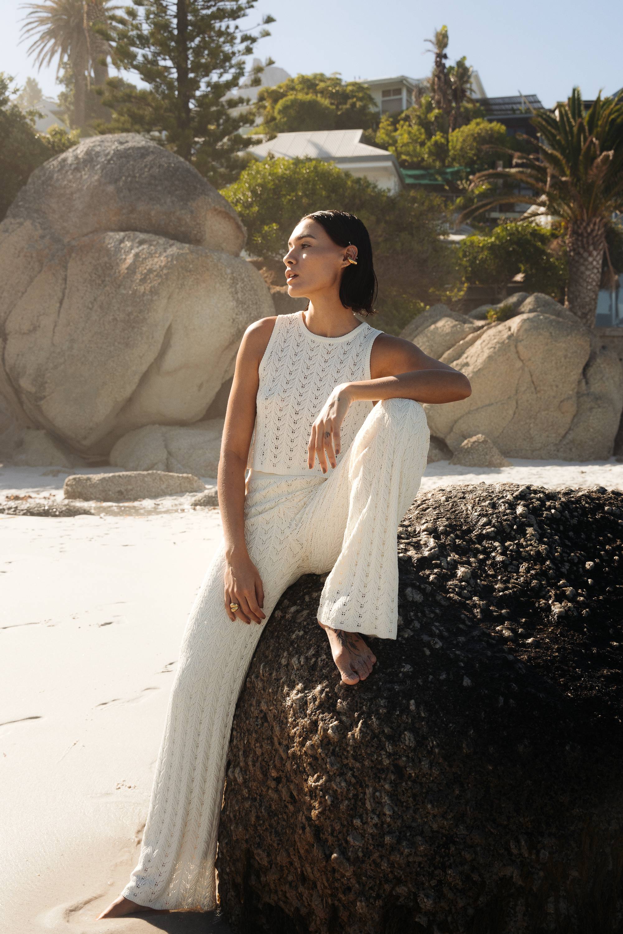 Woman in a white outfit sitting on a rock by a beach with palm trees and buildings in the background.